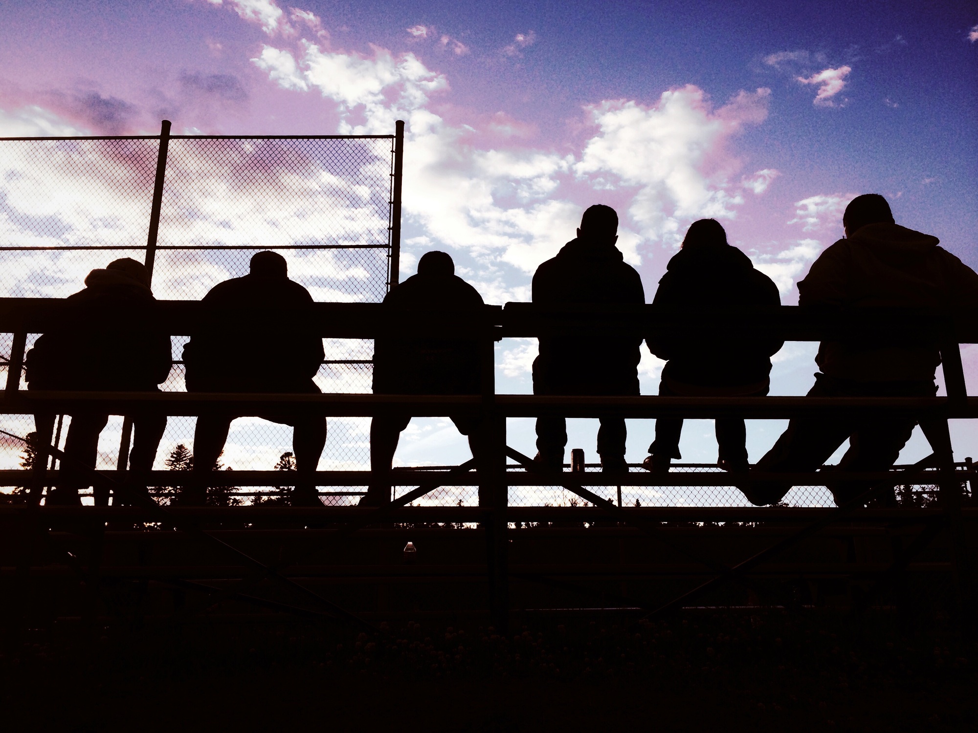 Silhouette from behind of people sitting on bleachers watching a ball game with evening purple sky.